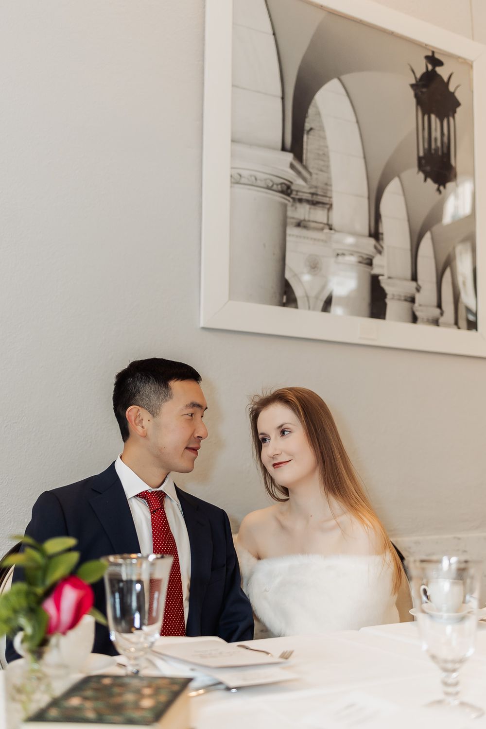 Newlywed couple sitting at wedding reception table, groom in navy suit with red tie, bride in white strapless gown.