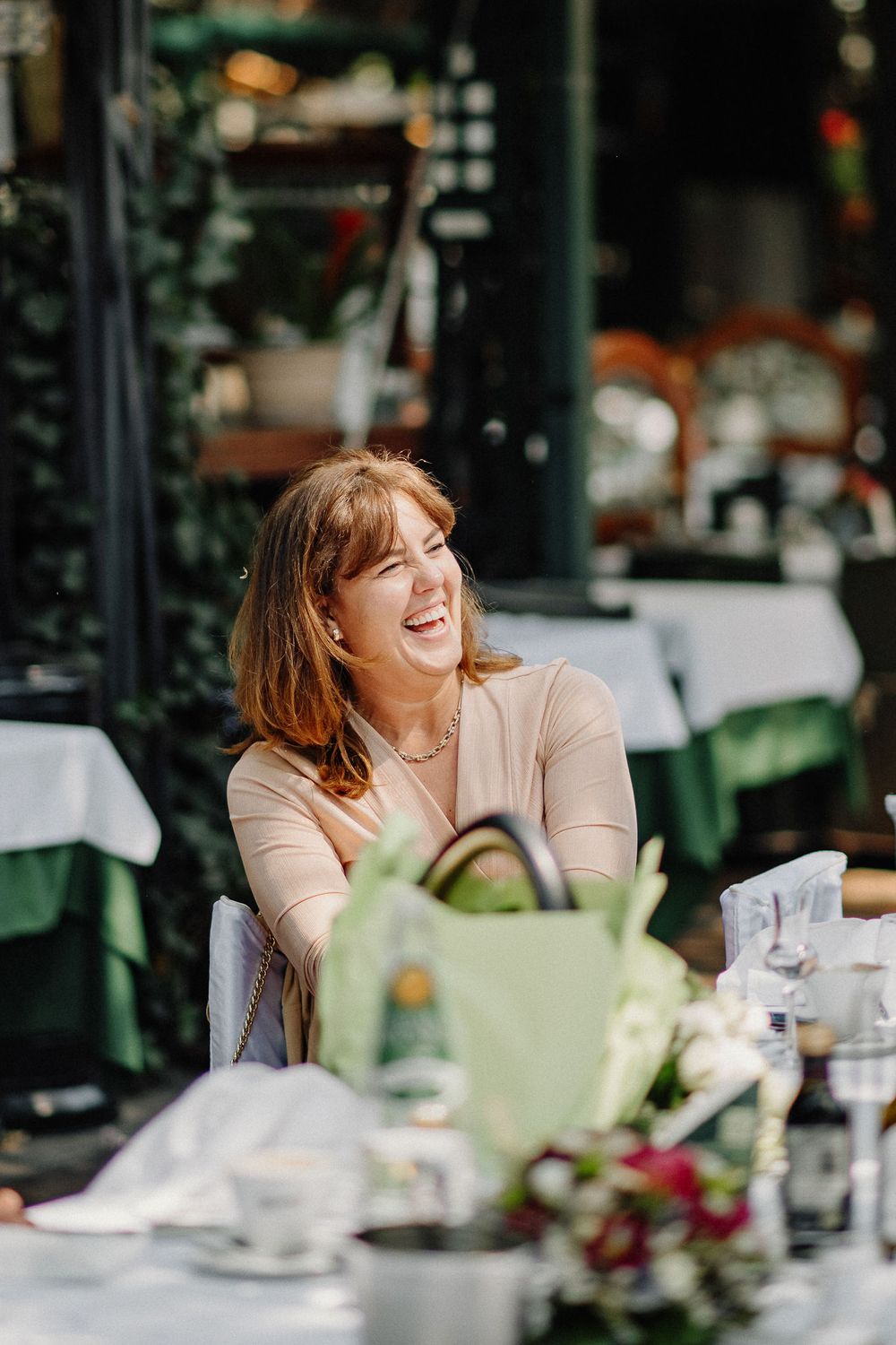 Woman enjoying afternoon tea and laughing at an outdoor cafe with table settings and greenery in the background.