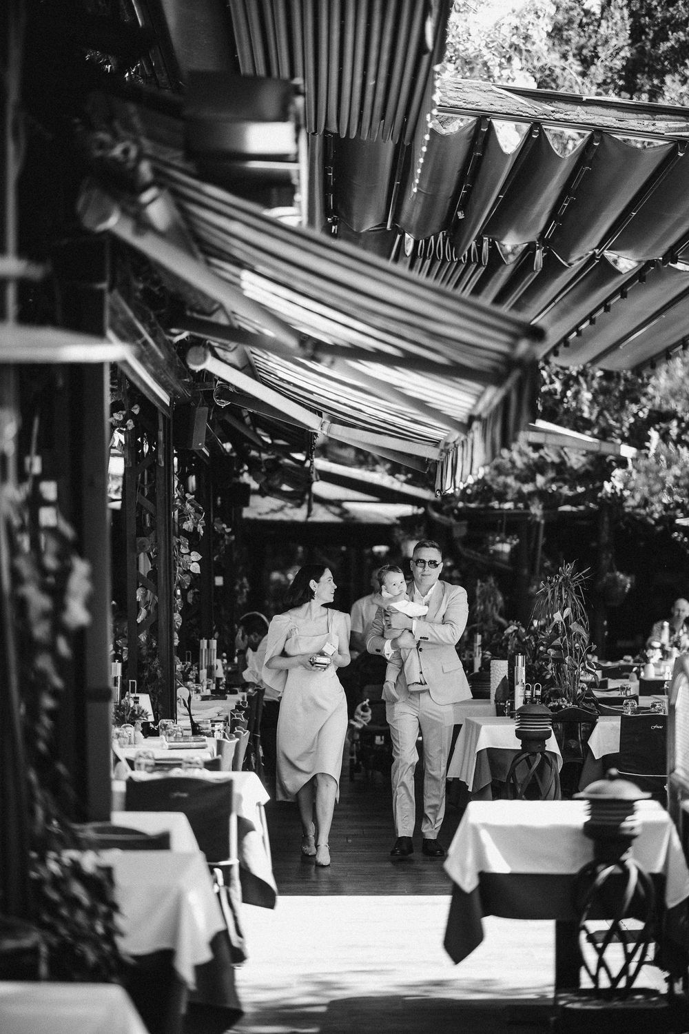 Restaurant service staff walk through an outdoor dining patio with cloth-covered tables beneath a striped awning.