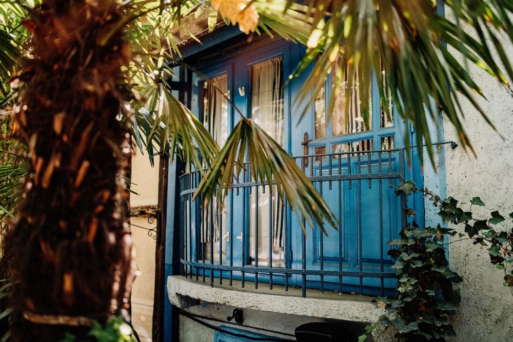 A vintage blue balcony with decorative ironwork framed by tropical palm leaves against a white building exterior.