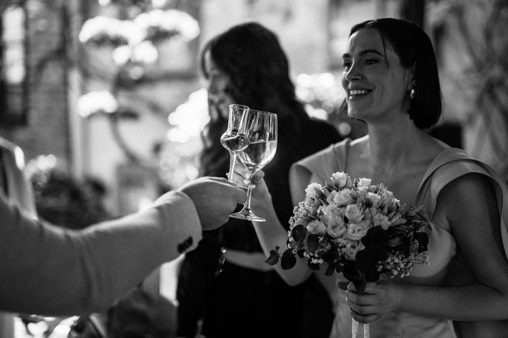 A monochrome photo of someone in formal attire holding a bouquet while toasting with a wine glass at a celebration.