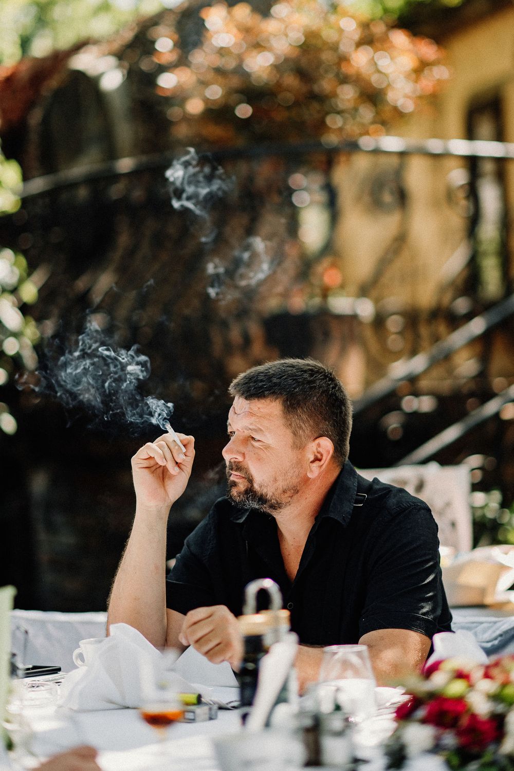 A person in a black shirt enjoying a cigarette while seated at an outdoor dining table on a sunny day.