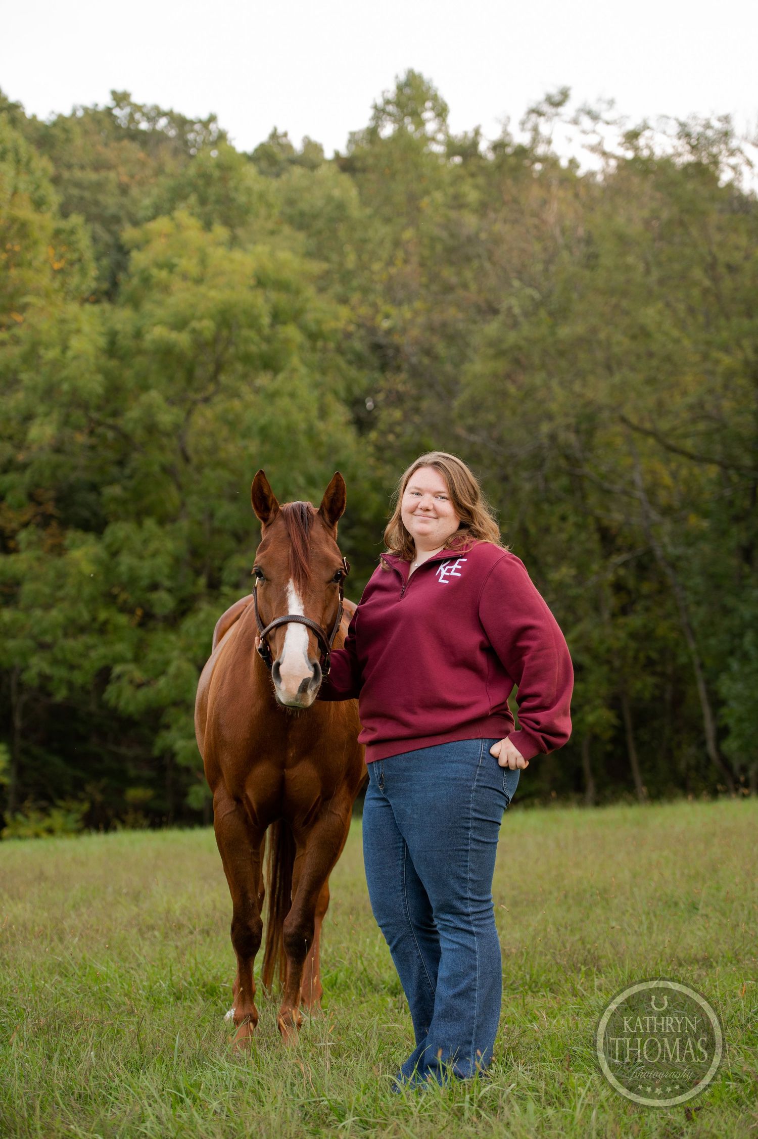 Chestnut horse and person in burgundy sweater standing in grassy field with autumn trees.