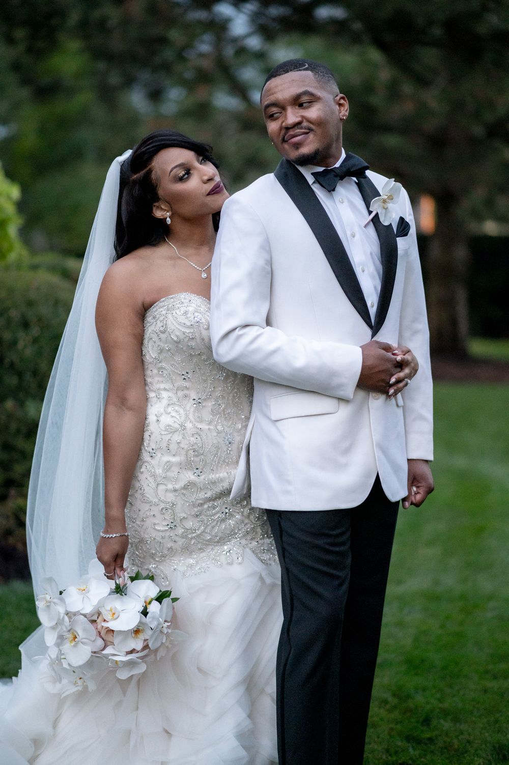 Elegant wedding couple poses in garden, bride in lace gown and groom in white tuxedo jacket.