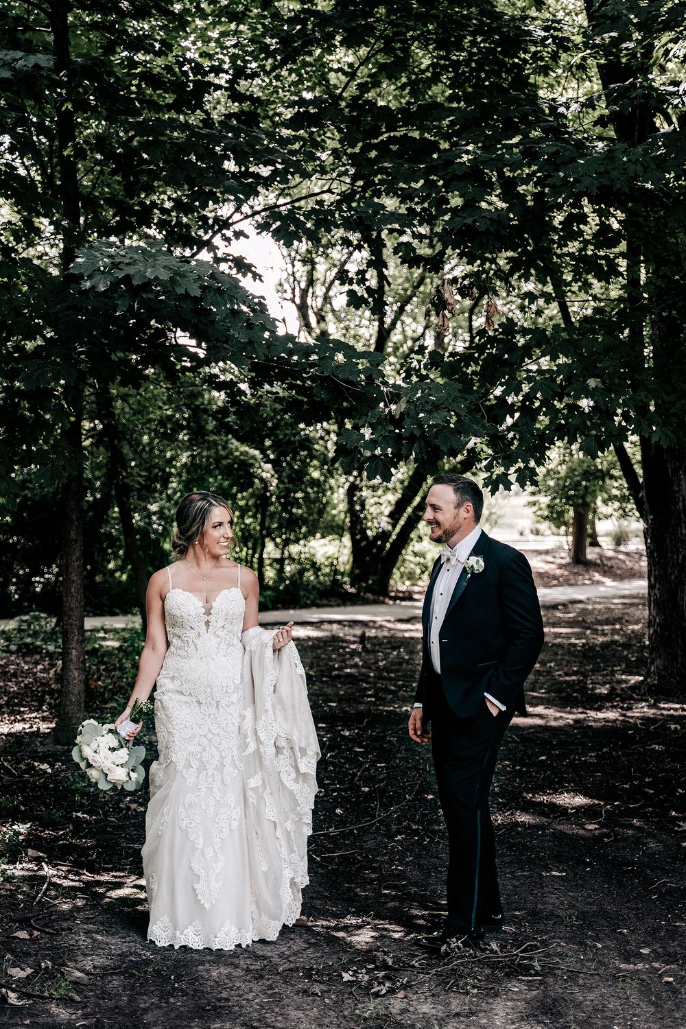 Wedding couple stands in wooded area, bride in lace gown holding white bouquet.