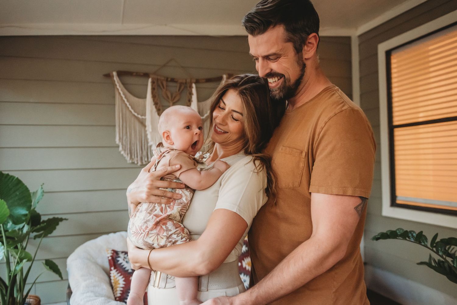 A family stands together on a porch with natural light coming through window blinds and a spiderweb decoration visible.