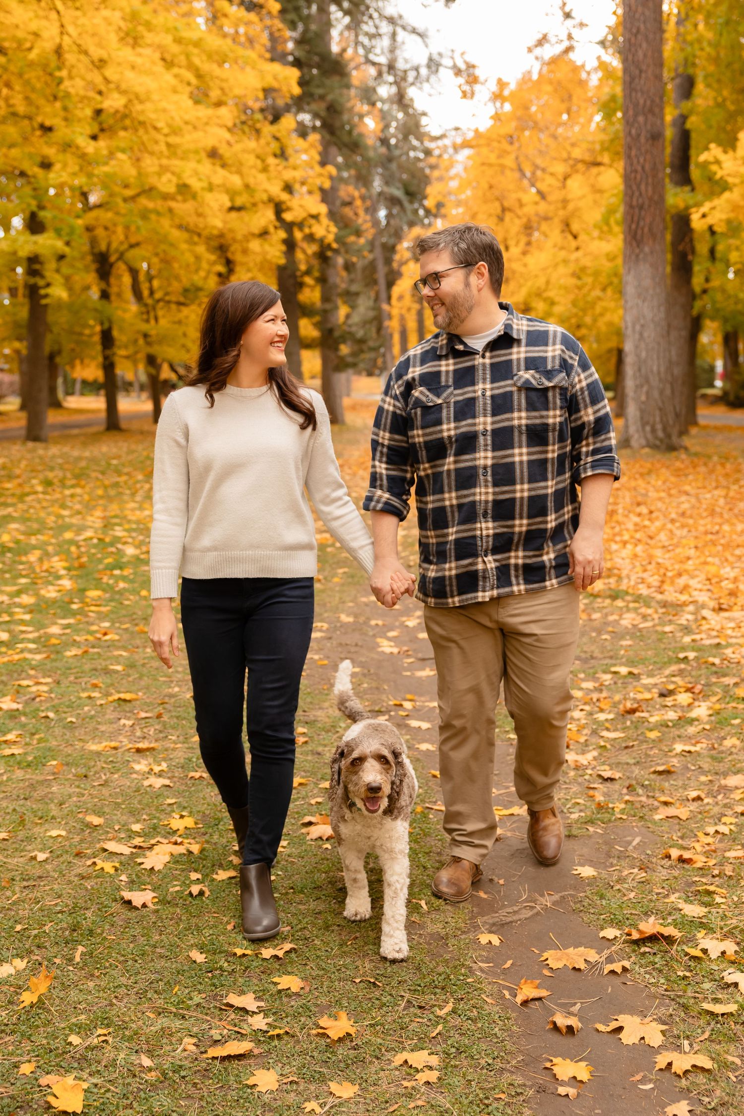 Couple walking dog on leaf-covered path through vibrant autumn forest. Spokane family photographer.