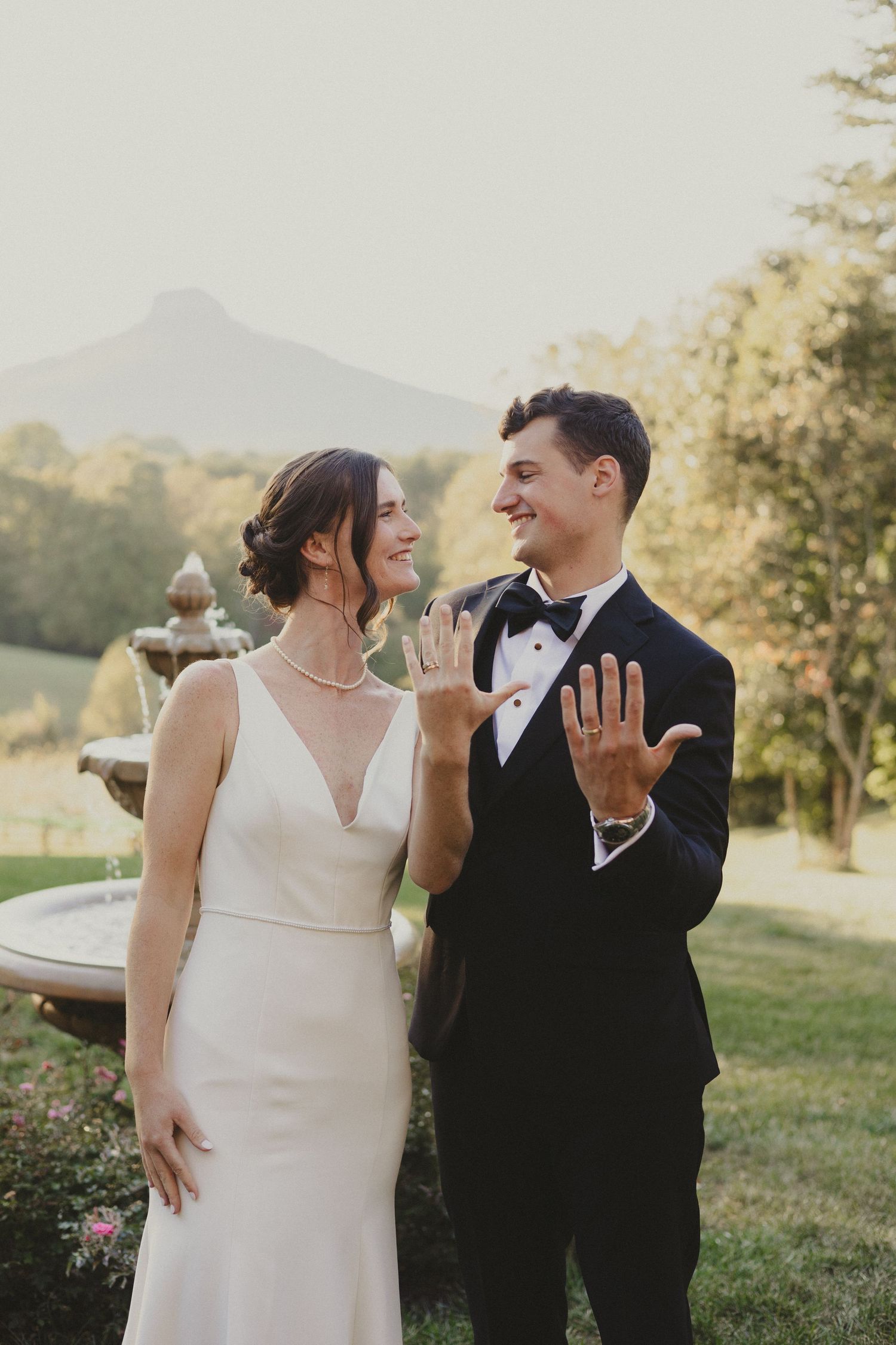 Newlyweds showing off rings in garden setting with mountain backdrop.