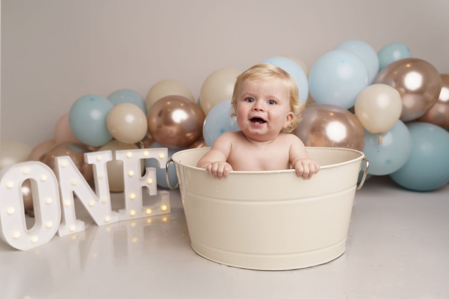A one year old baby sits in a cream colored bucket surrounded by blue and gold balloons and ONE letter decorations.