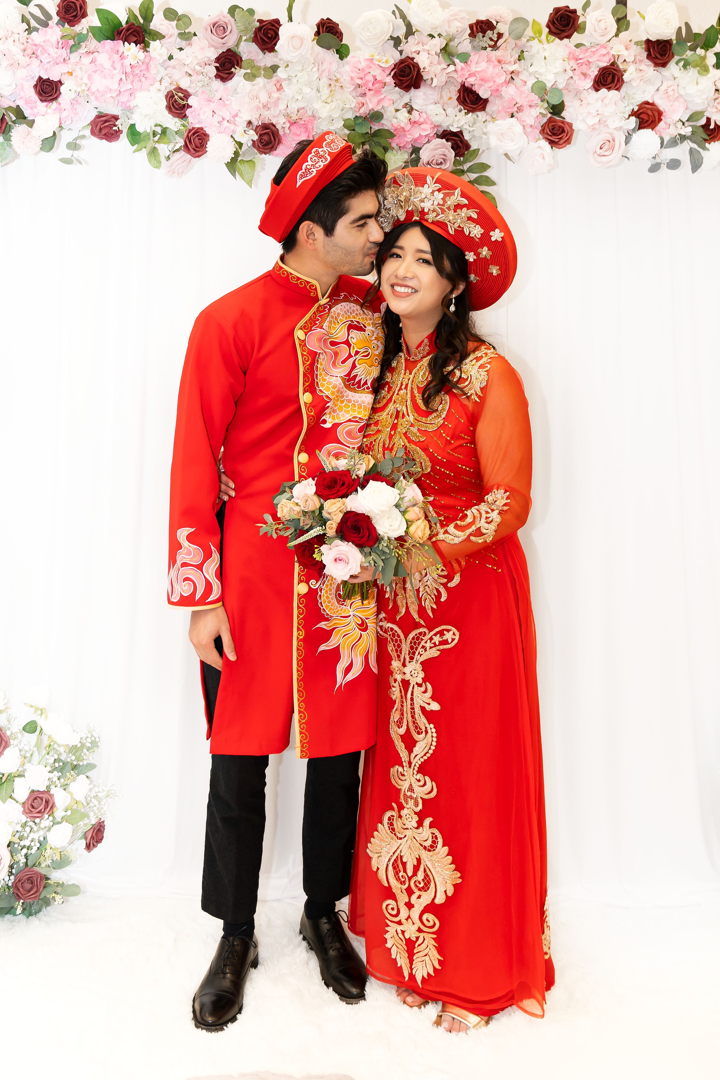 Red traditional Vietnamese wedding attire with floral backdrop featuring matching red ao dai and headdresses.