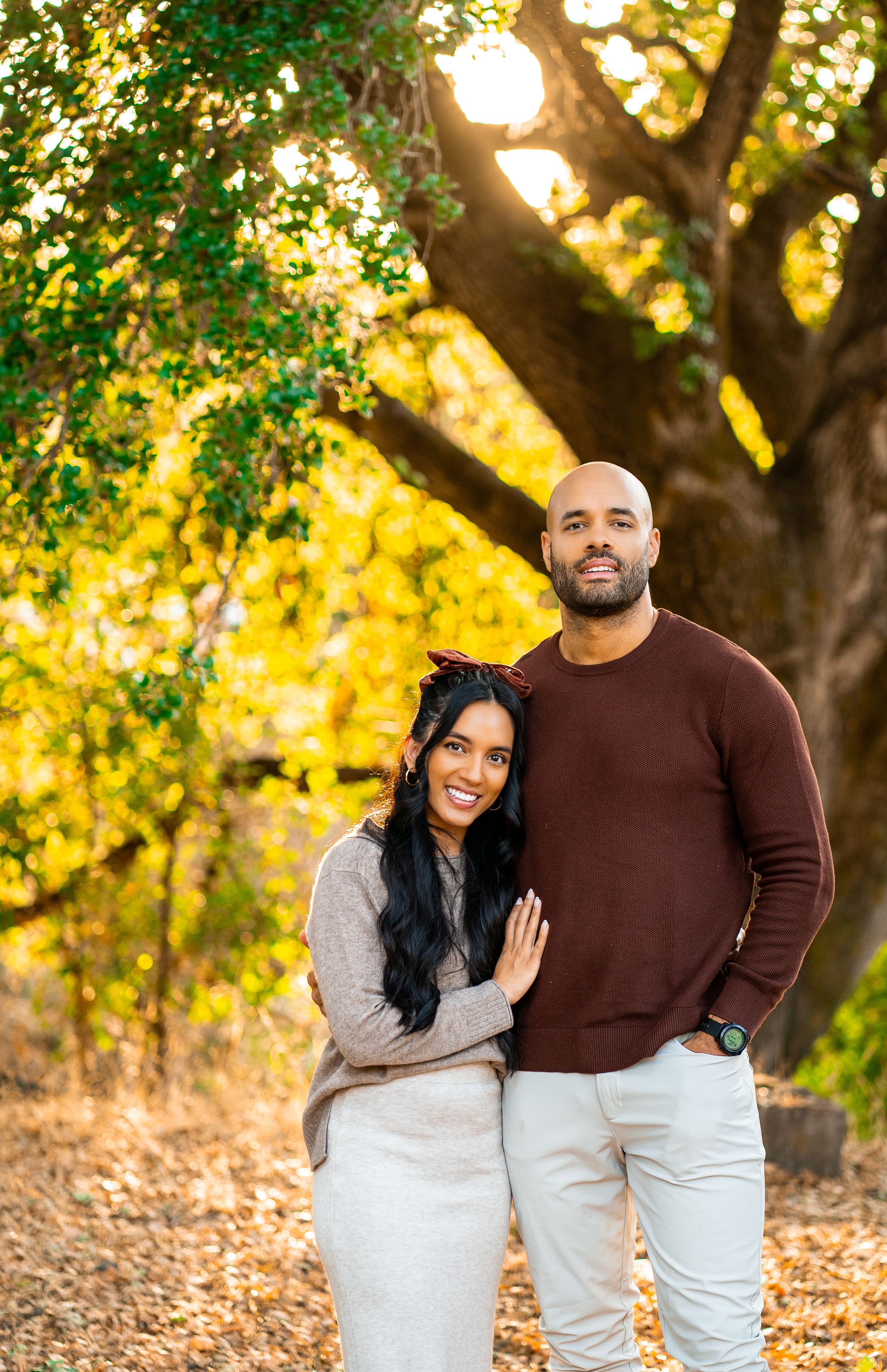 A couple embraces in a romantic autumn setting with golden sunlight filtering through trees behind them.