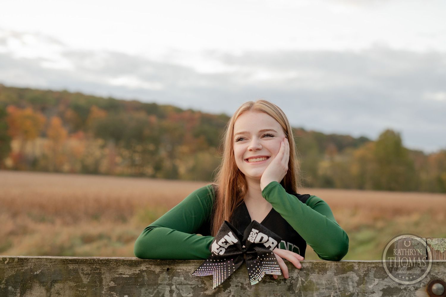 Student in green sweater leans on wooden fence in autumn field setting.