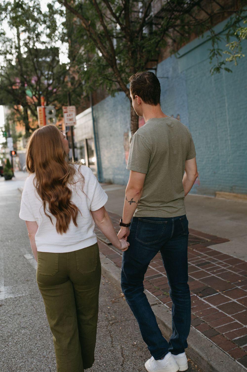 Couple walking hand in hand along urban sidewalk with blue wall and trees.