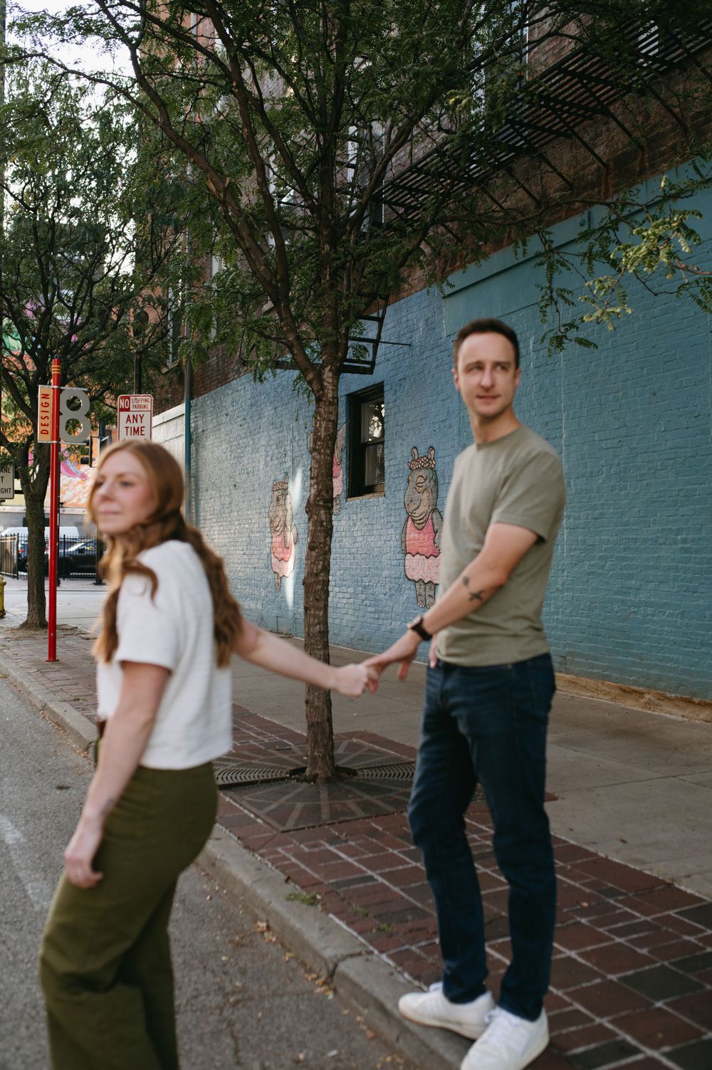 Romantic couple walking on city sidewalk beside blue wall and trees.