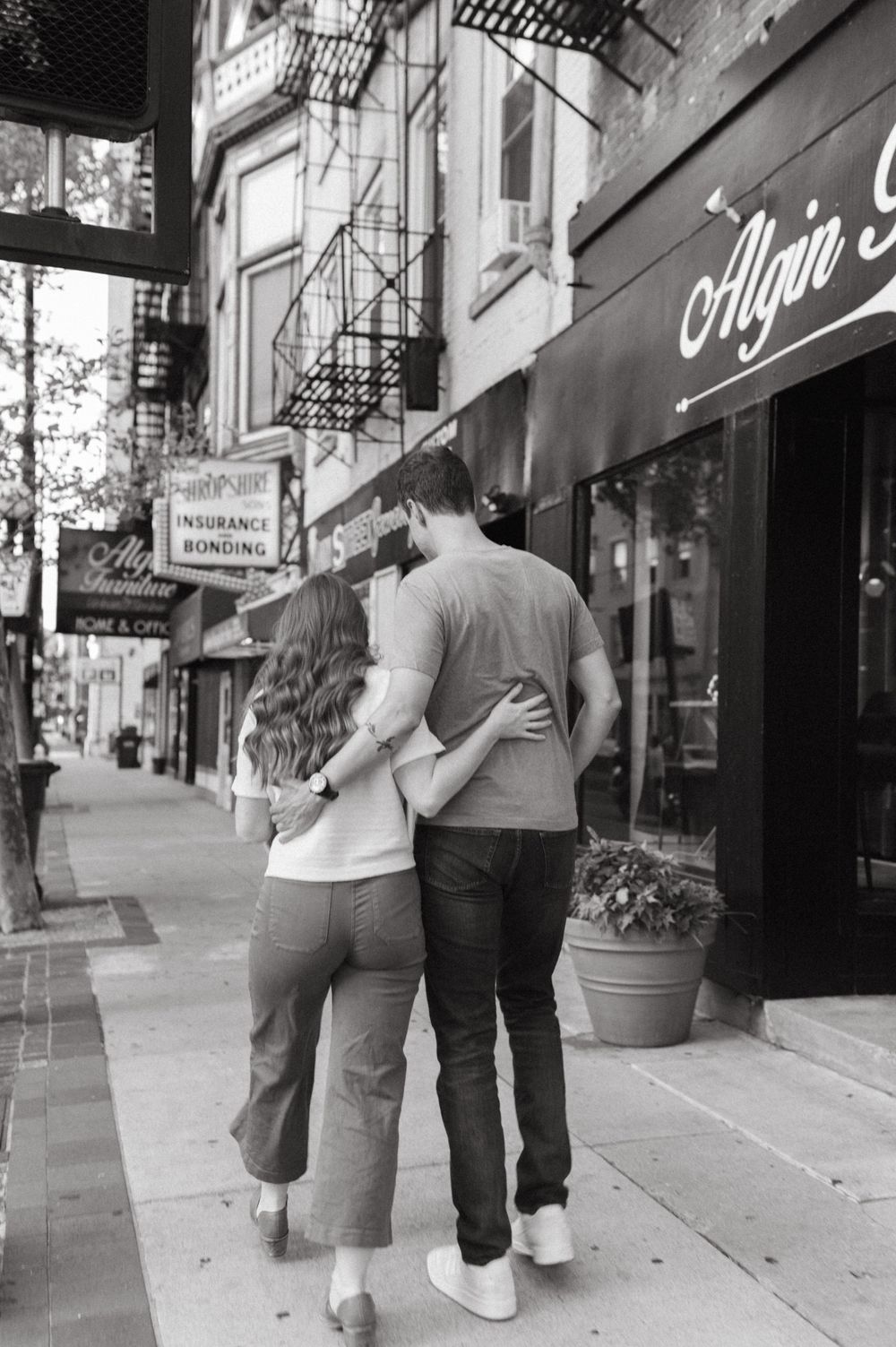 A couple embraces while walking down a city sidewalk in black and white.