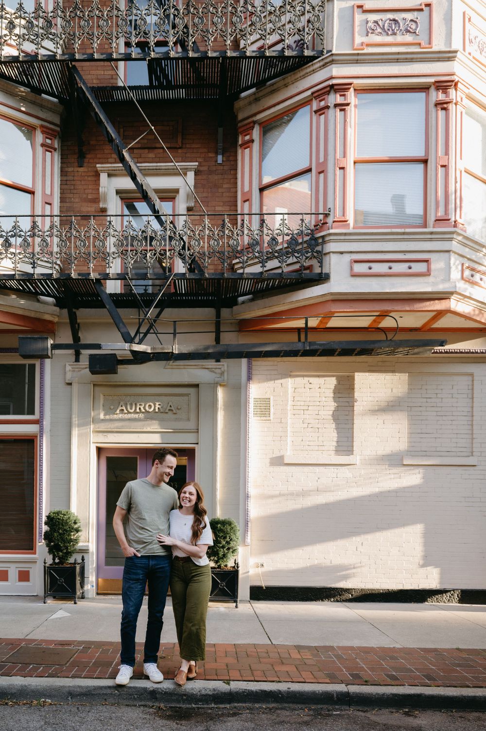 Couple posing in front of historic brick building with ornate iron fire escapes.