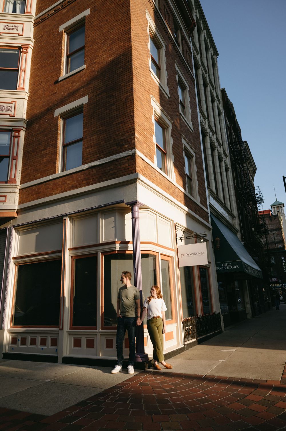 A couple stands outside a historic brick building with large windows on a city corner.