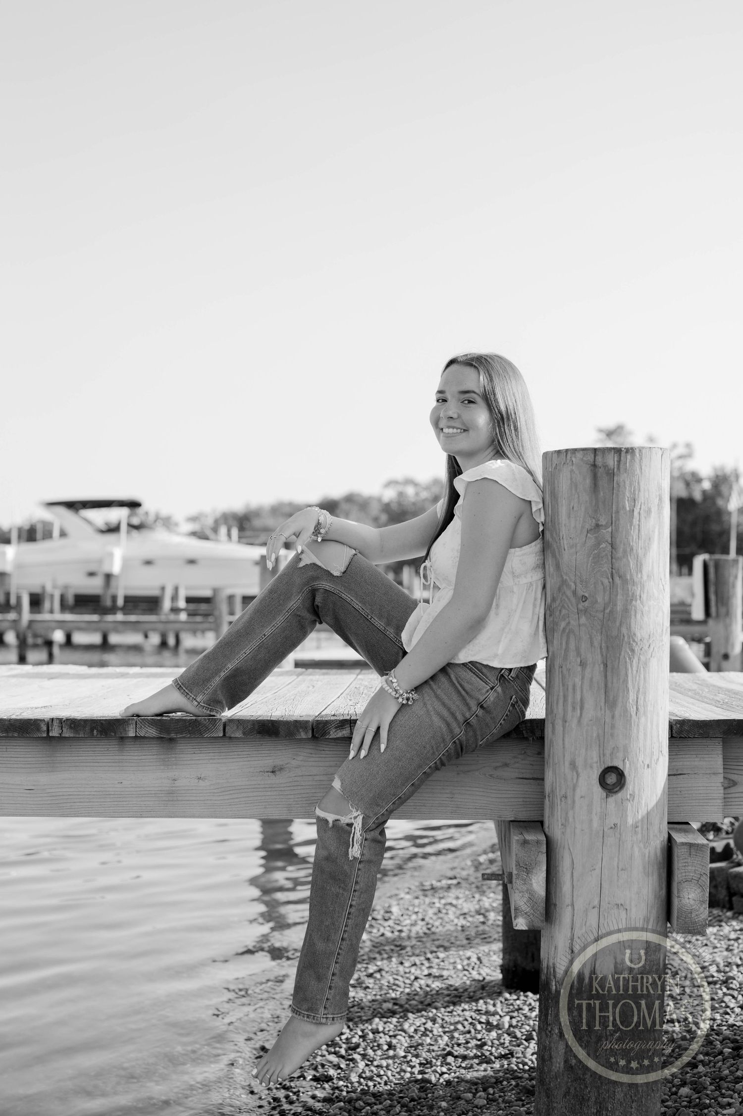 Black and white portrait of someone sitting on a dock piling by the water.