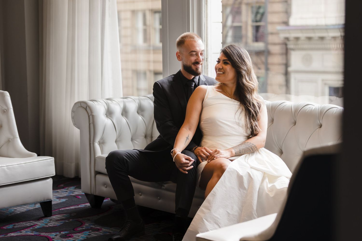 A couple shares an intimate moment on an elegant tufted gray couch by a window in a luxurious indoor setting.