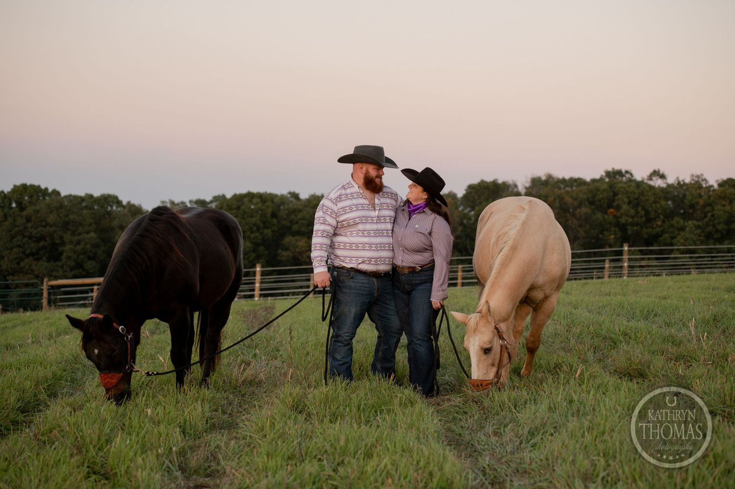 Two horses graze while couple stand together in a fenced pasture during sunset.