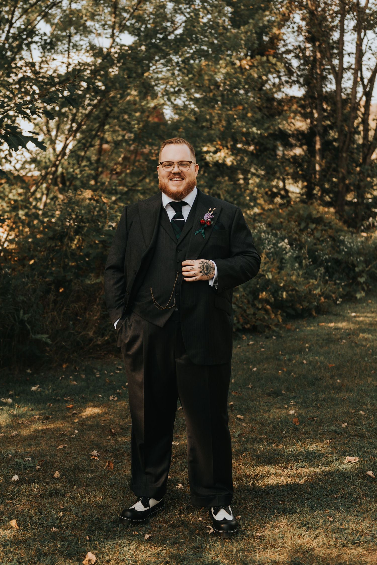 Man in black suit and dress shoes holds coffee cup while standing outdoors among autumn trees in warm lighting.