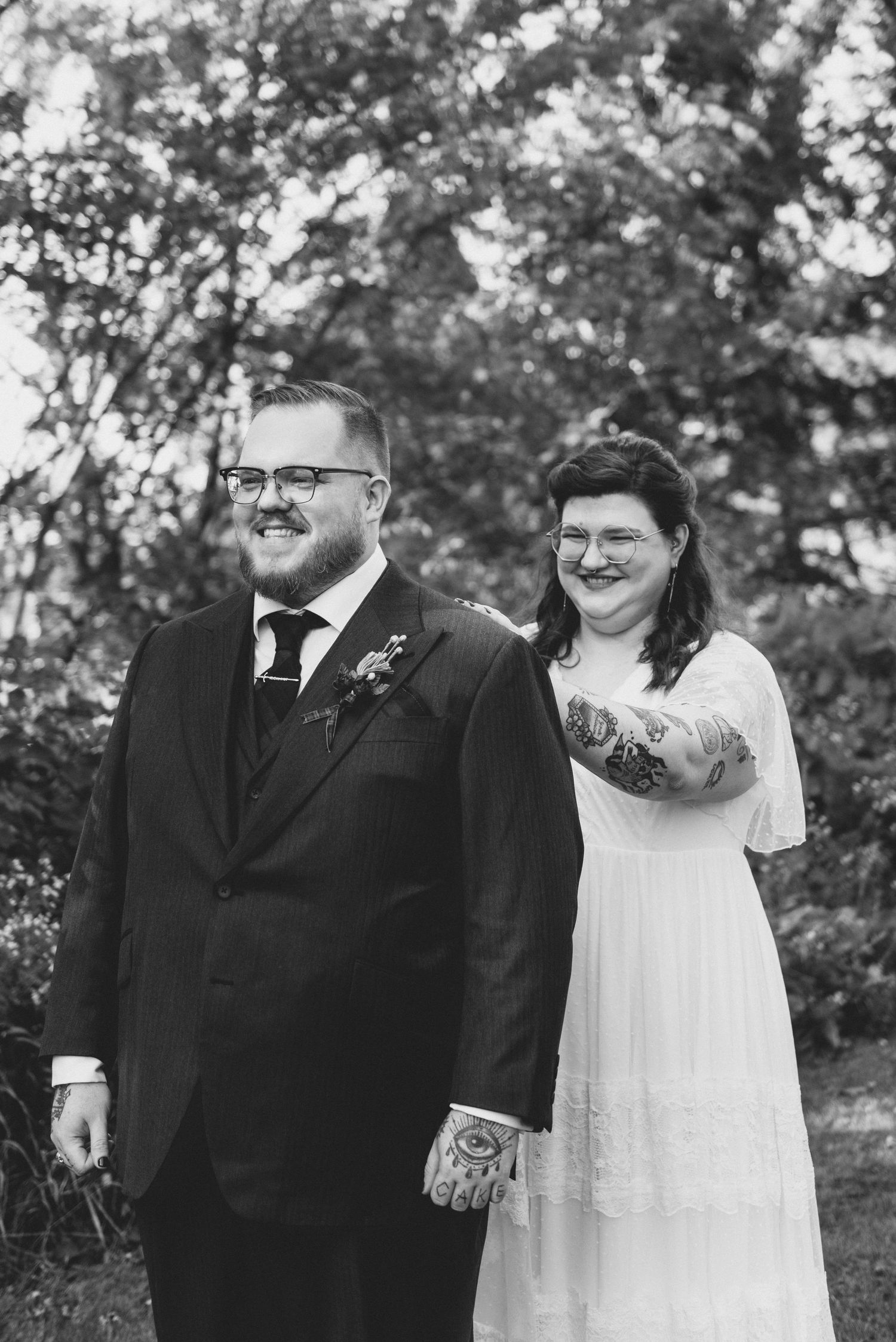 A black and white photograph of a happy couple on their wedding day, standing outdoors with trees in the background.