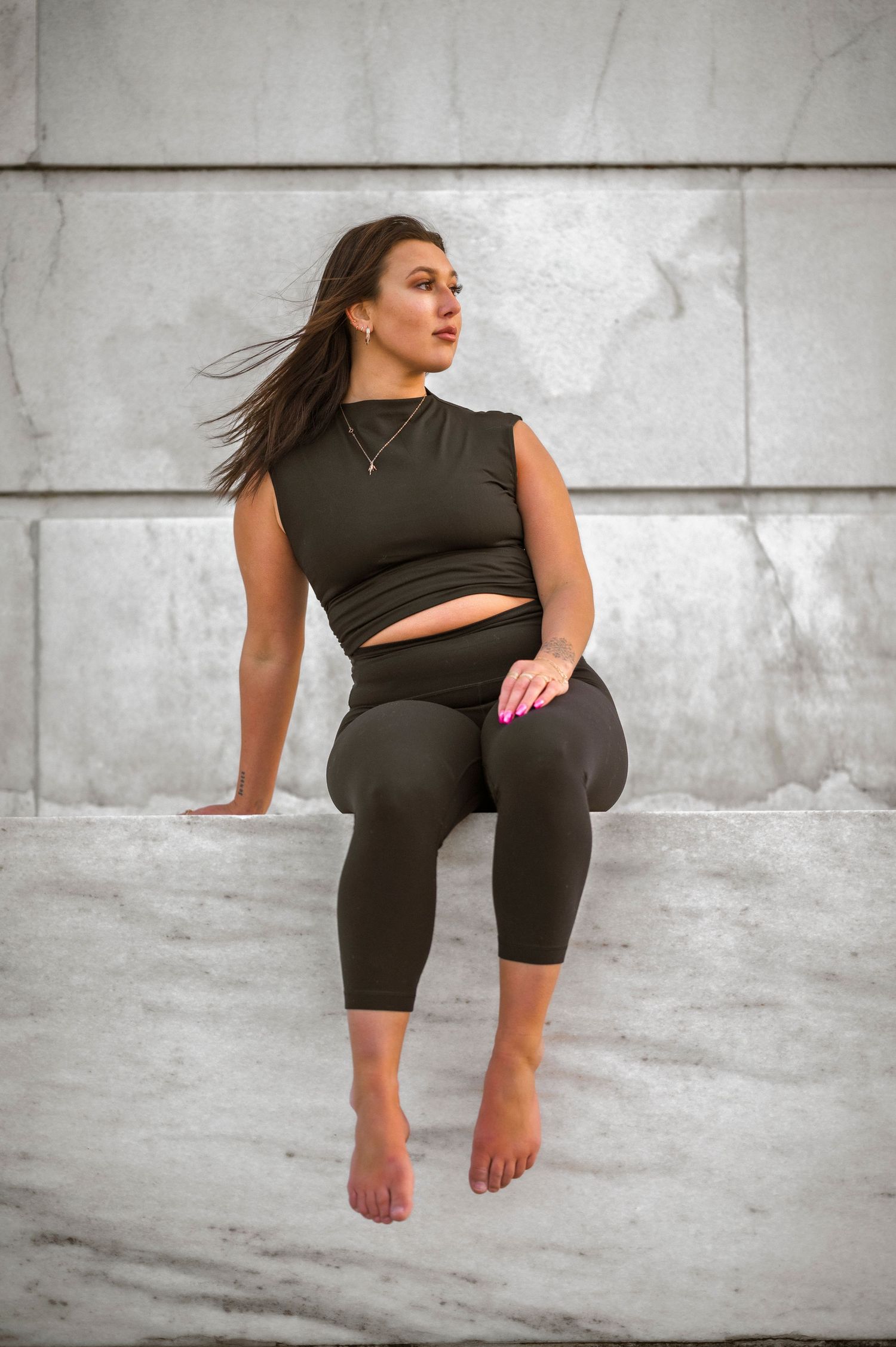 Athletic woman in black crop top and leggings poses with wind-blown hair against concrete wall.