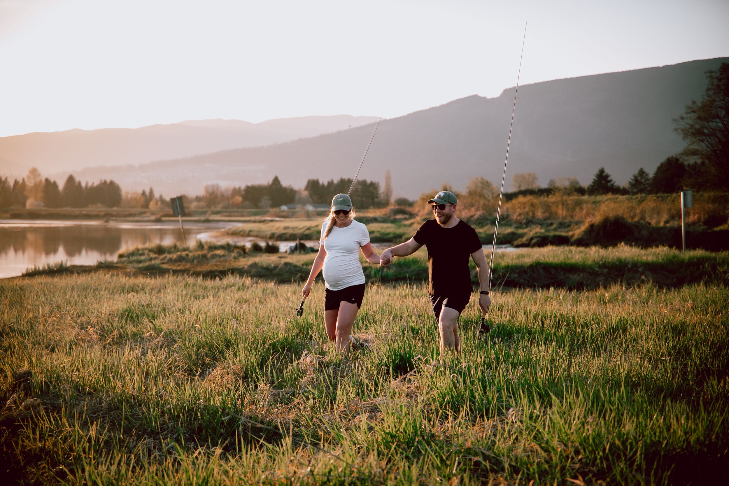 Two people walking through tall grass during golden hour with mountains in background.