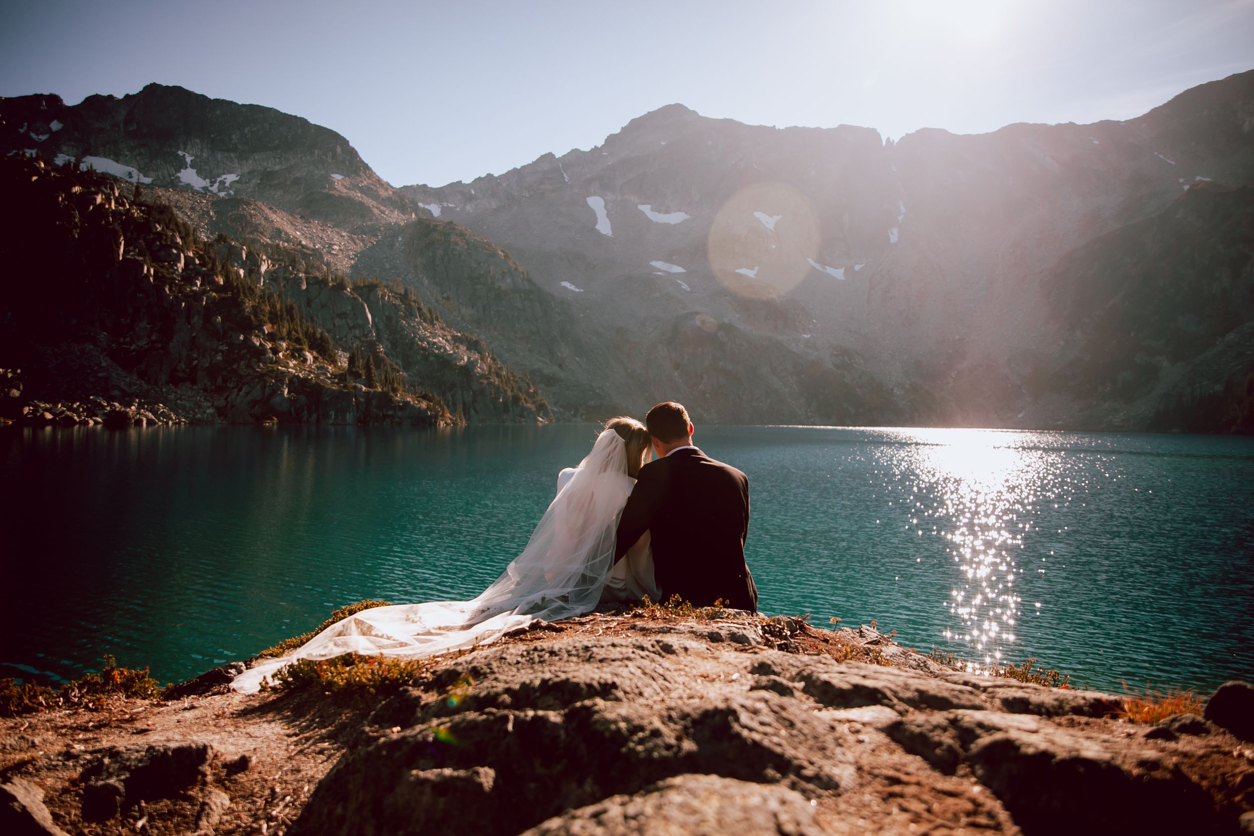 Couple in wedding attire sitting on cliff edge overlooking turquoise mountain lake.