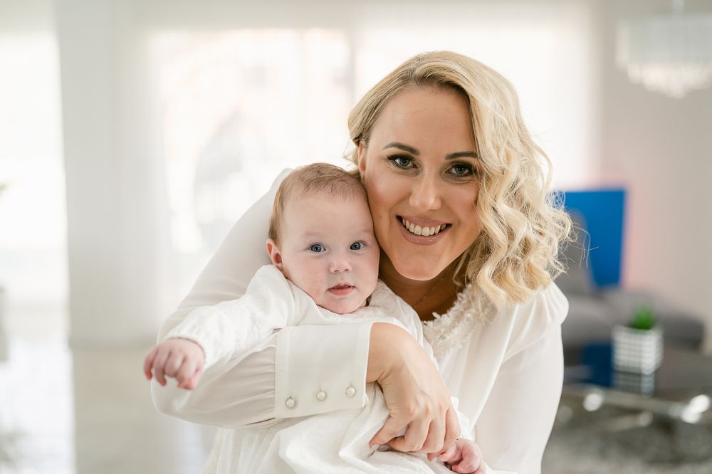 Mother and baby in white outfits pose together in bright indoor setting.