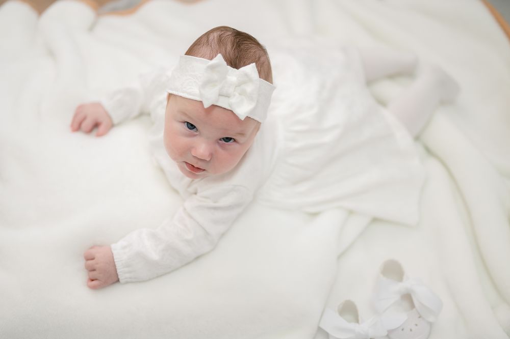 A baby wearing a white headband lies on soft white blankets in a gentle pose.
