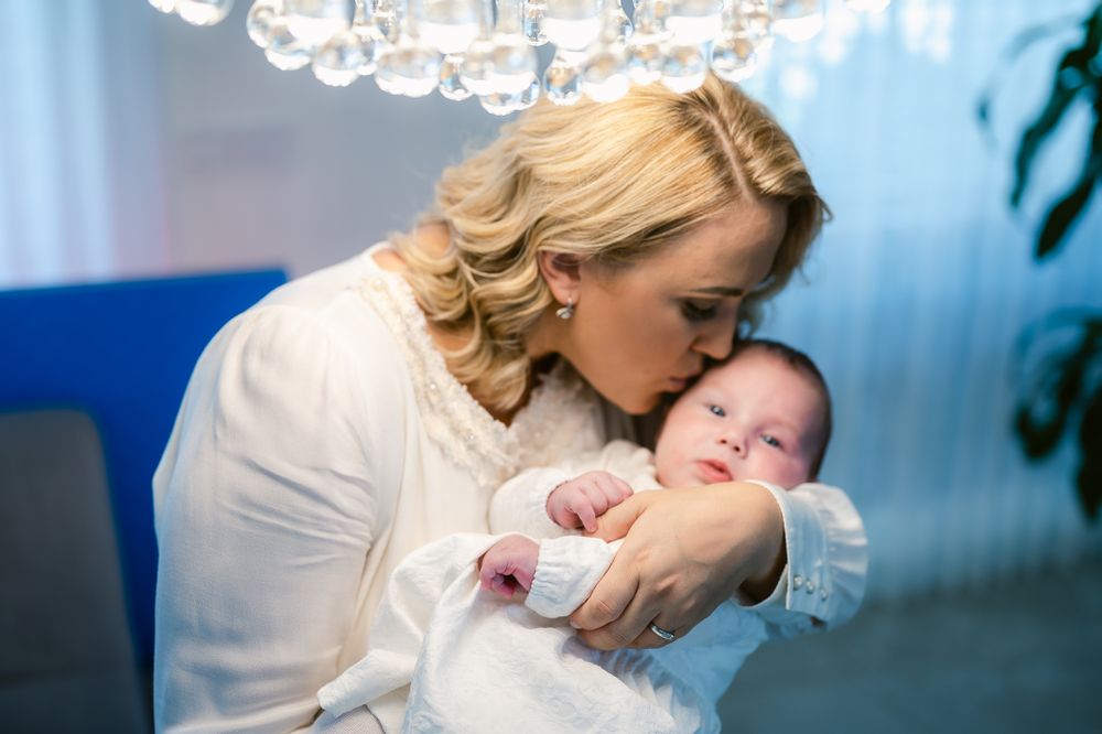 A tender moment shows a maternal embrace with a sleeping infant wearing white under soft chandelier lighting.