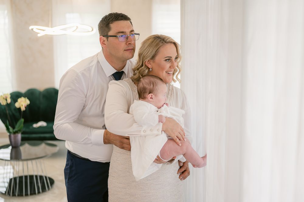 A couple in formal white attire holds their baby in a bright room with floral decor.