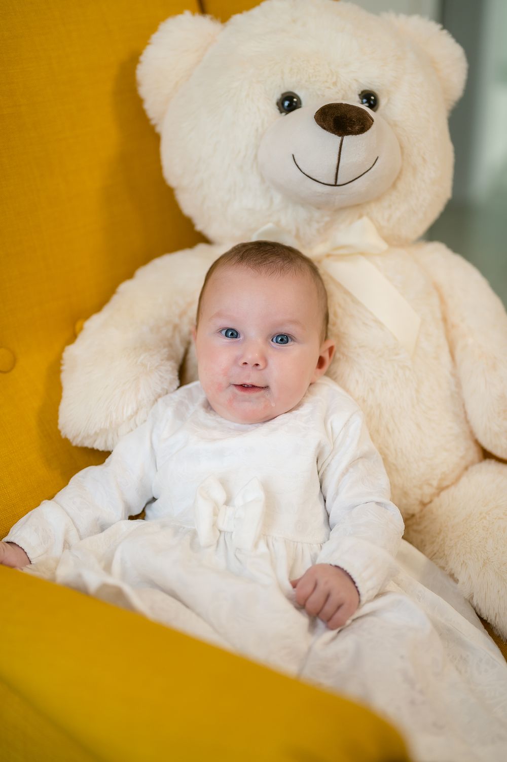 A small baby in white clothing sits against a large cream-colored teddy bear on a yellow background.
