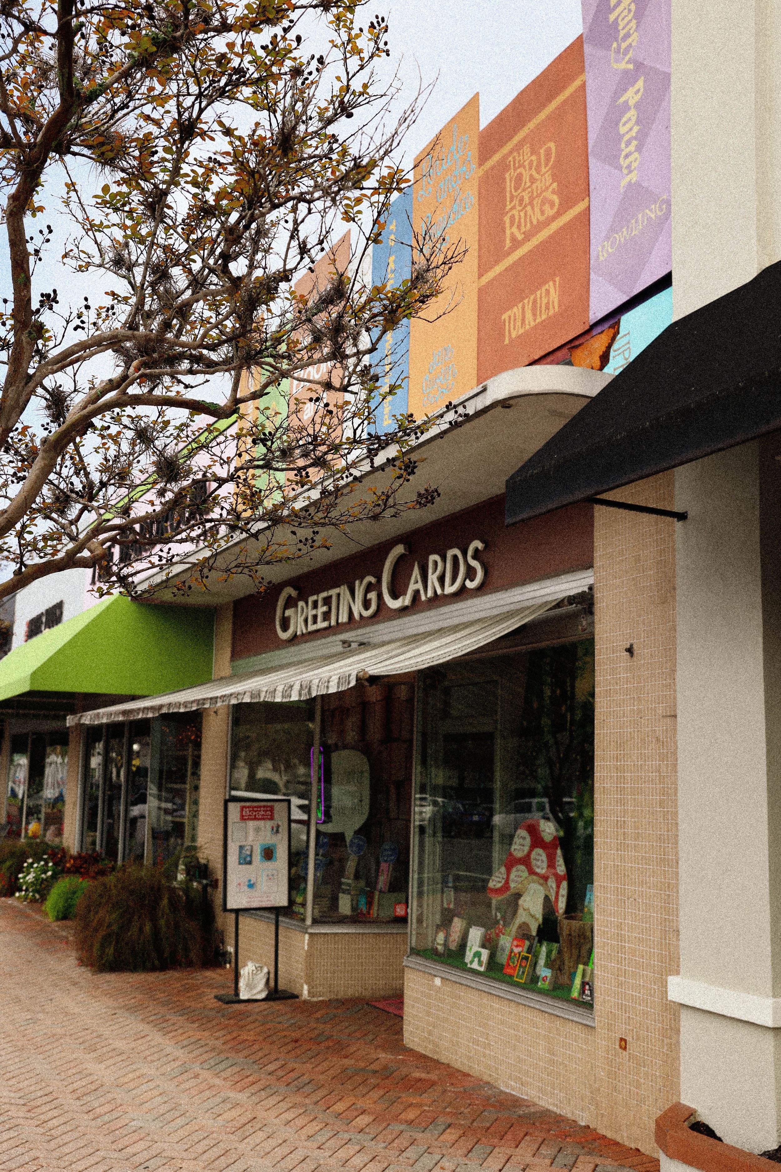 Exterior view of a greeting card store with brick sidewalk and colorful signage along a shopping district storefront.