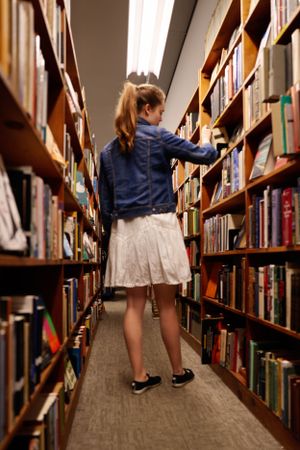 A student in a denim jacket and white skirt browses books in a library aisle between tall wooden shelves.