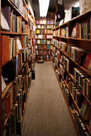 Narrow aisle between tall wooden bookshelves filled with books in a library or bookstore with warm lighting.