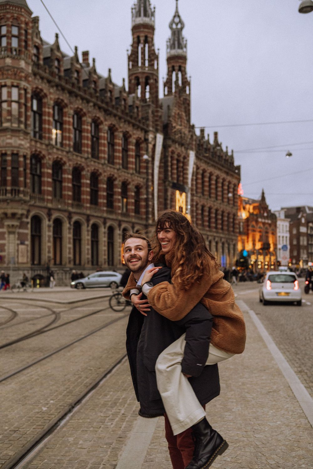 Smiling couple enjoying a piggyback ride on a tram-lined cobblestone street in front of a grand red-brick Gothic building at dusk.