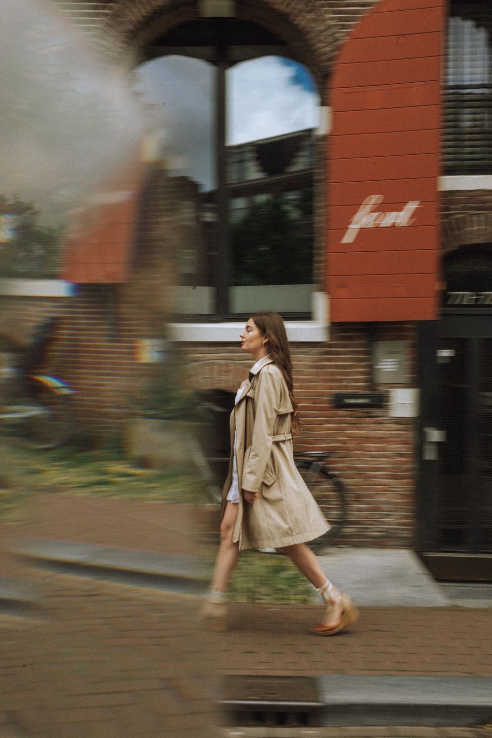 Motion blur sequence of person in beige coat walking past brick buildings.