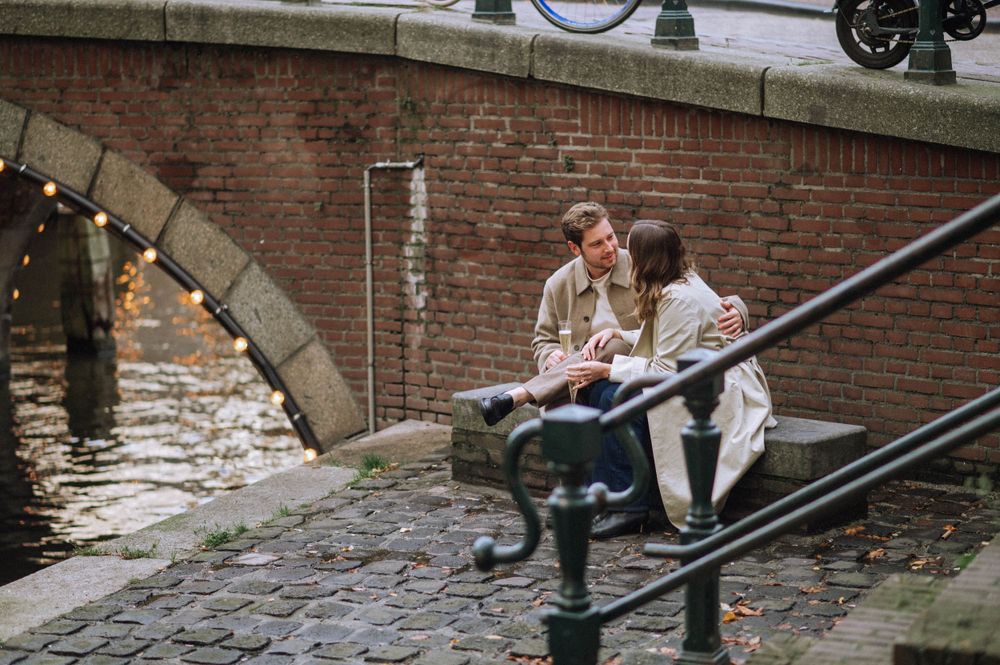 A couple sits together on stone steps by a canal with string lights overhead.