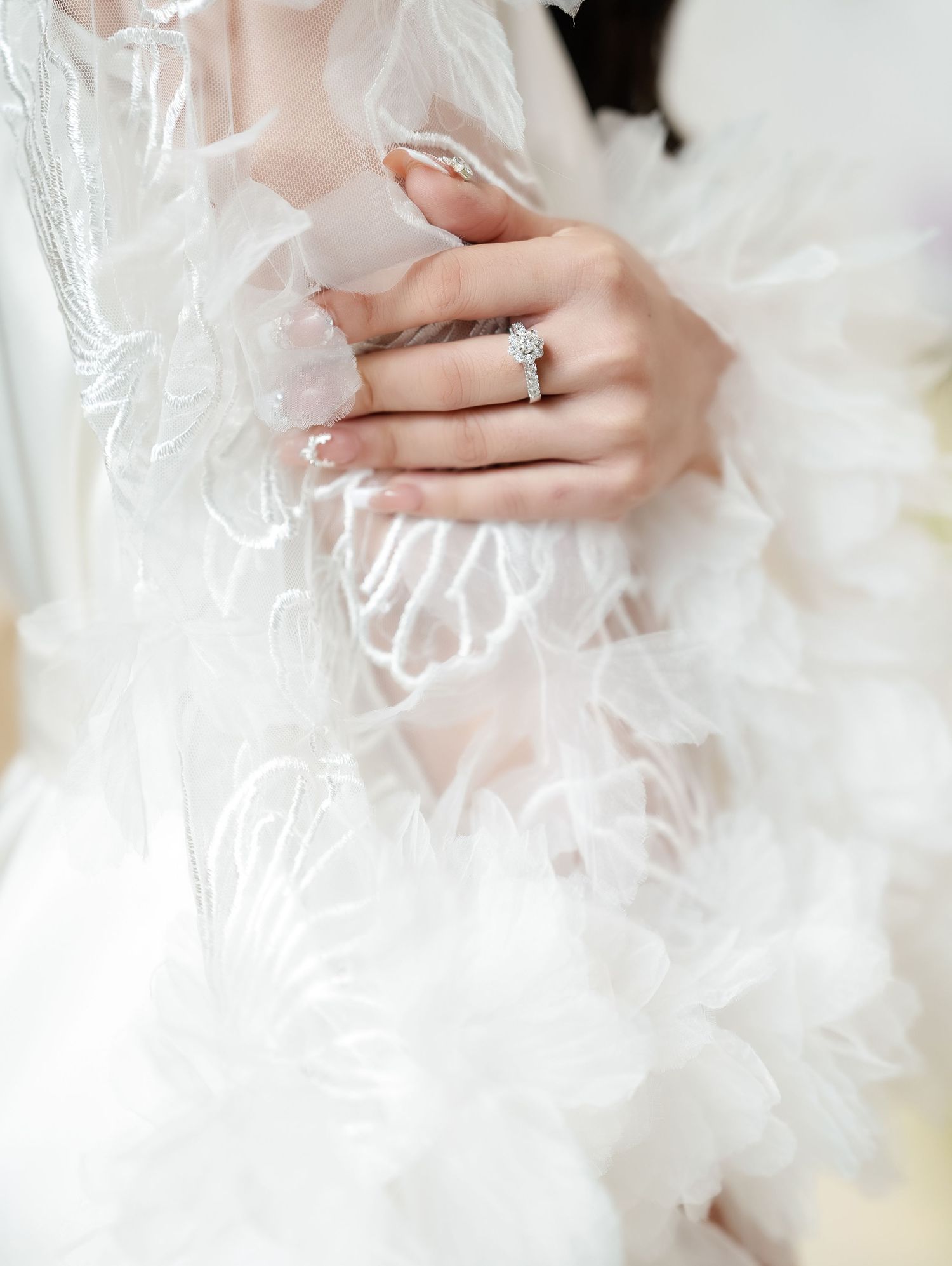 Close up of an elegant diamond engagement ring on a delicate hand against ethereal lace fabric details.