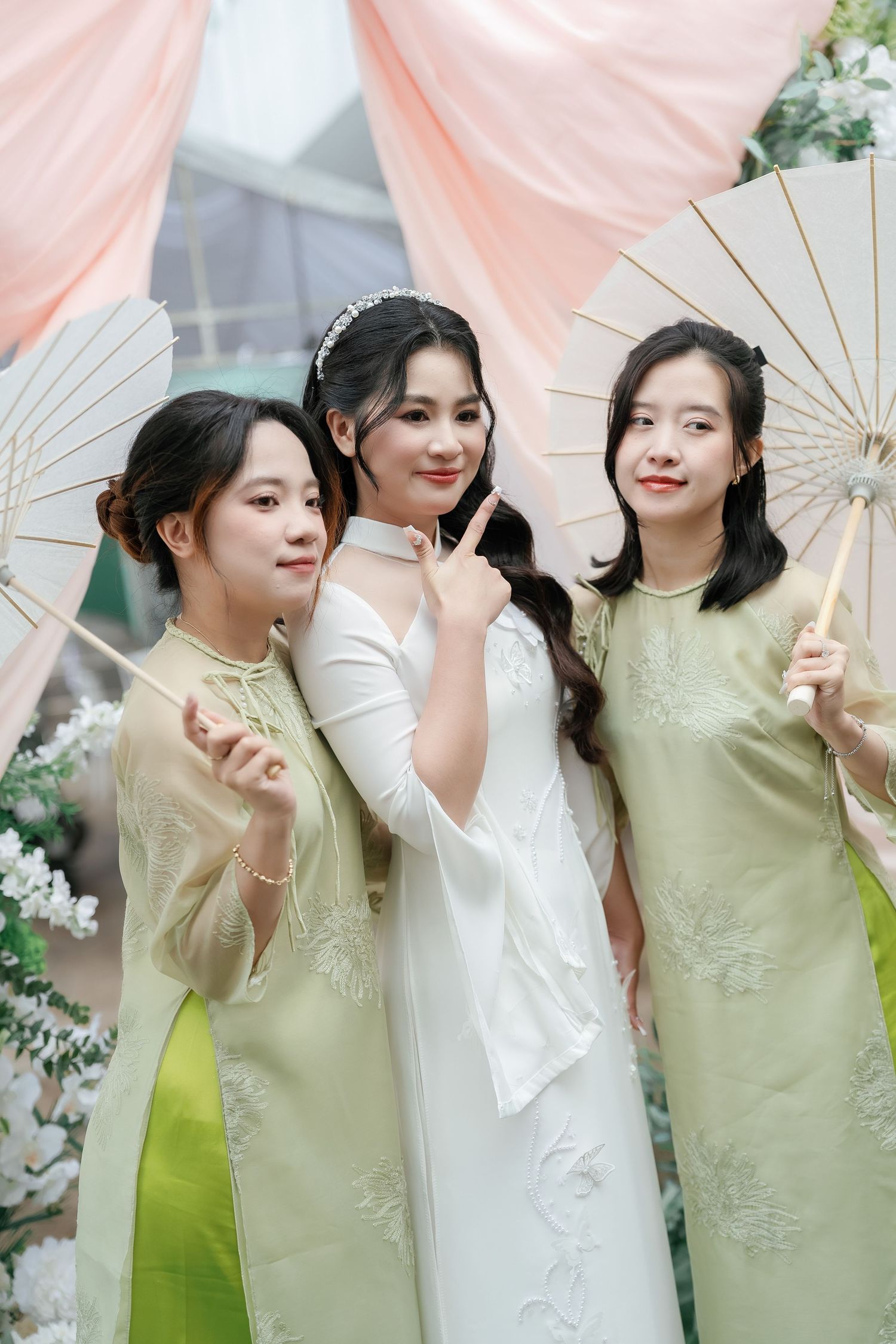 Three people in elegant attire pose with decorative parasols at a wedding with peach drapes and floral decor.