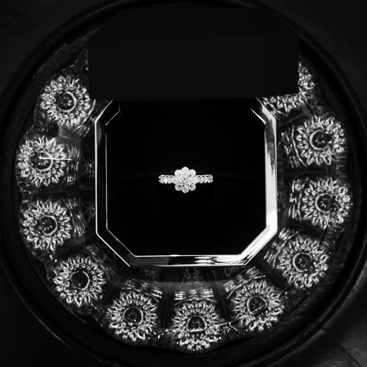 Black and white photo of a diamond engagement ring surrounded by ornate fractal-like designs in a circular pattern.