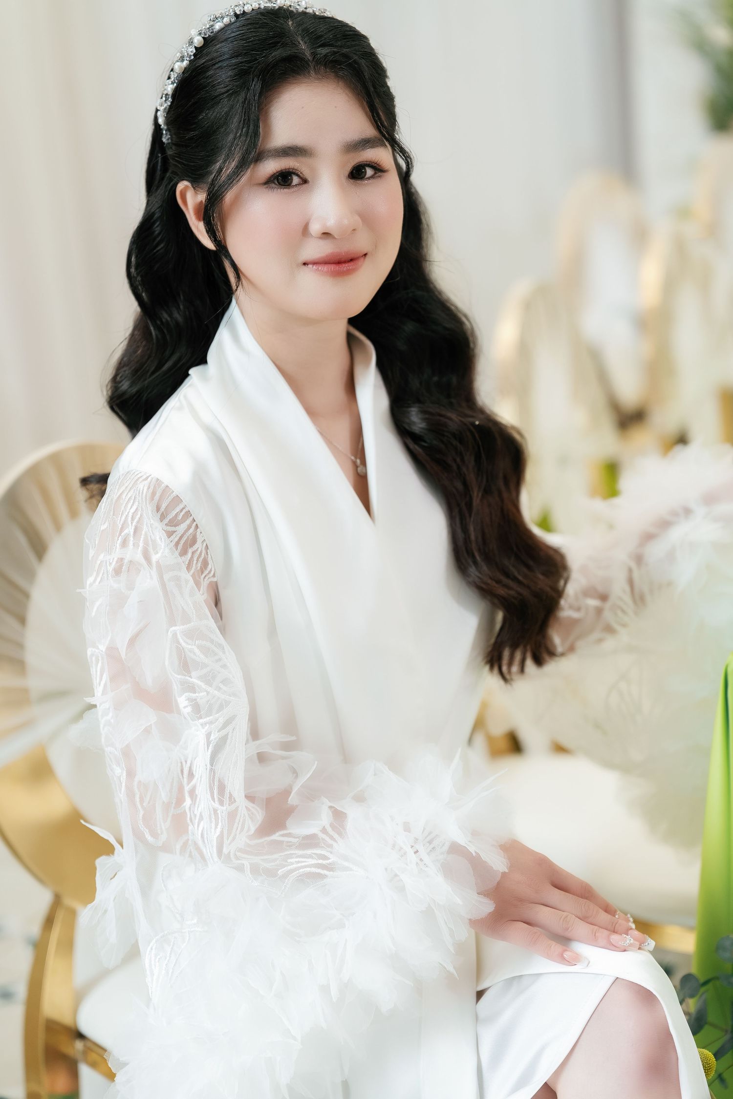 A person in a white lace robe sits elegantly on a chair in a bright, softly lit setting.