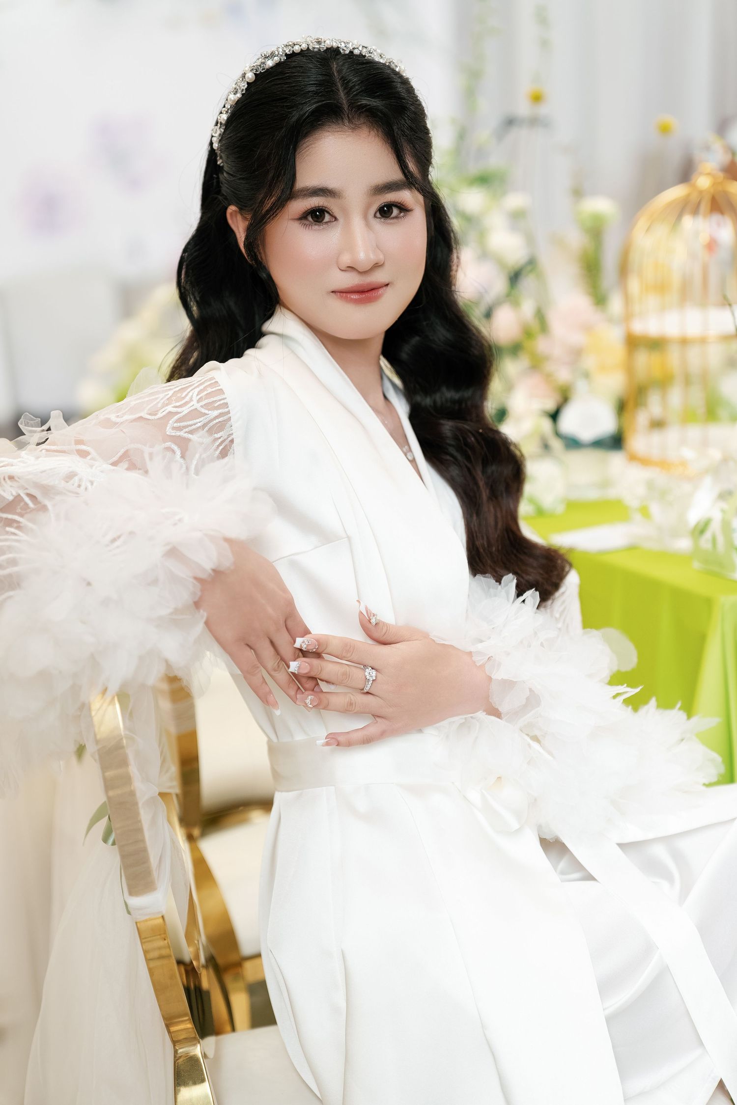 Elegant figure in white silk robe with feather accents poses against floral backdrop at luxury event setting.