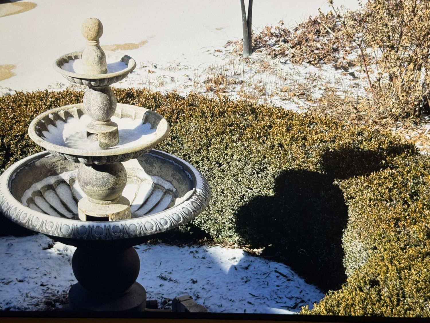 Stone garden fountain casting shadows on snowy ground with trimmed hedges.