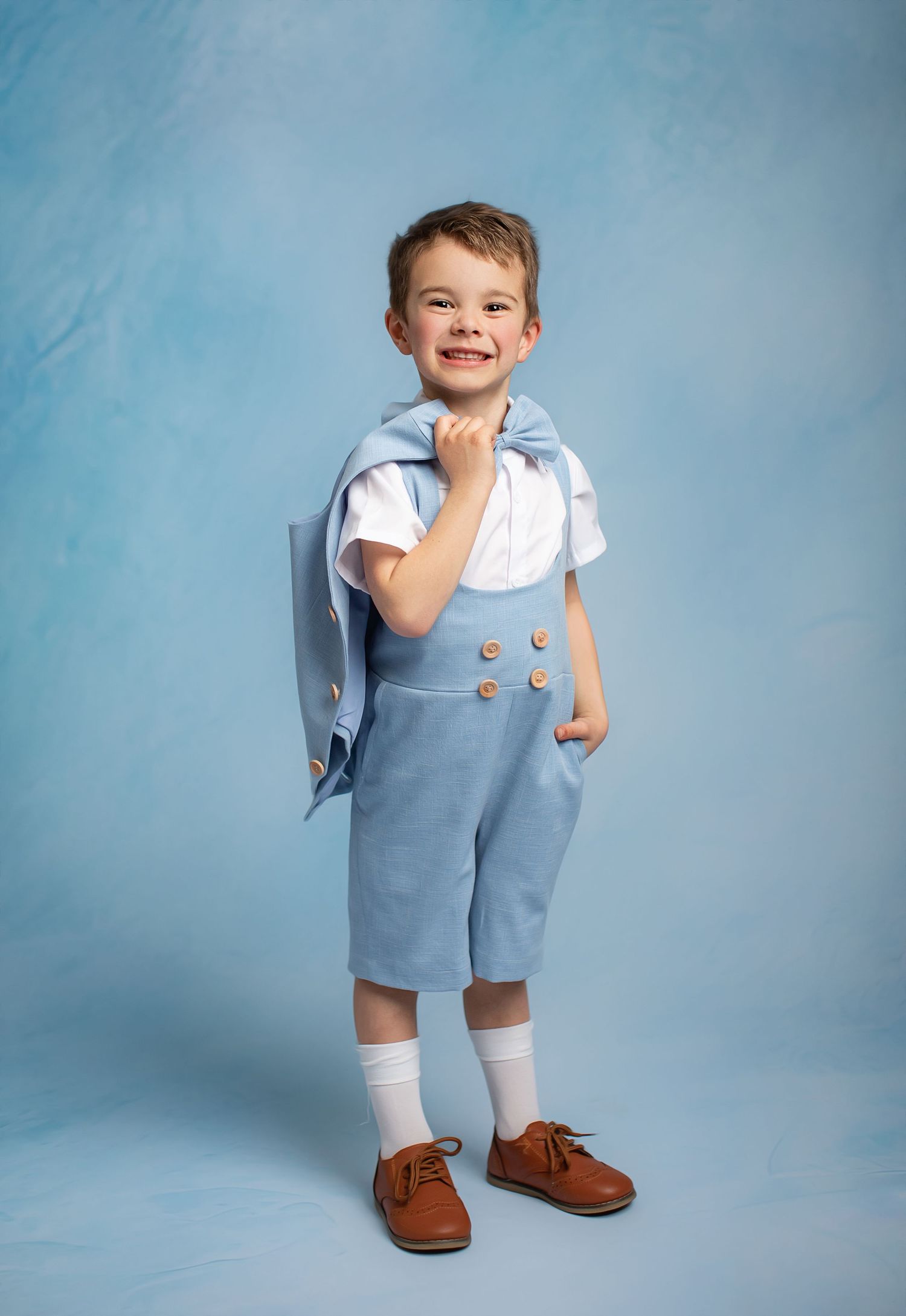 A small figure in a light blue romper with white shirt and brown shoes poses against a blue studio backdrop.