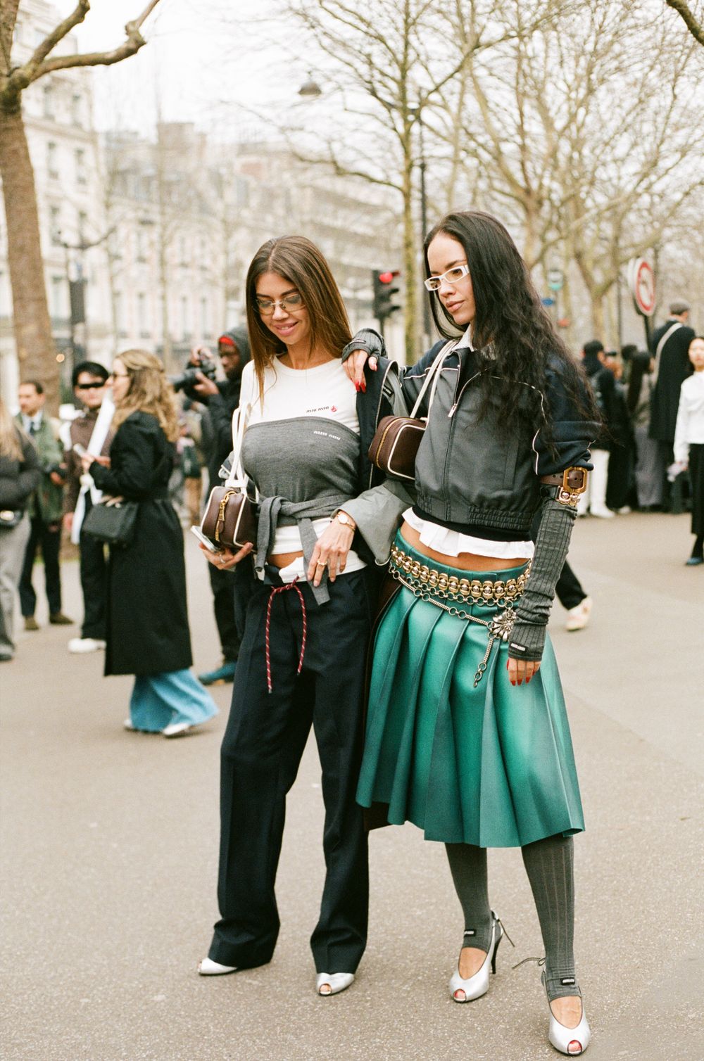 Two fashionable women in street style outfits pose together during fashion week wearing contrasting skirt and pant looks.