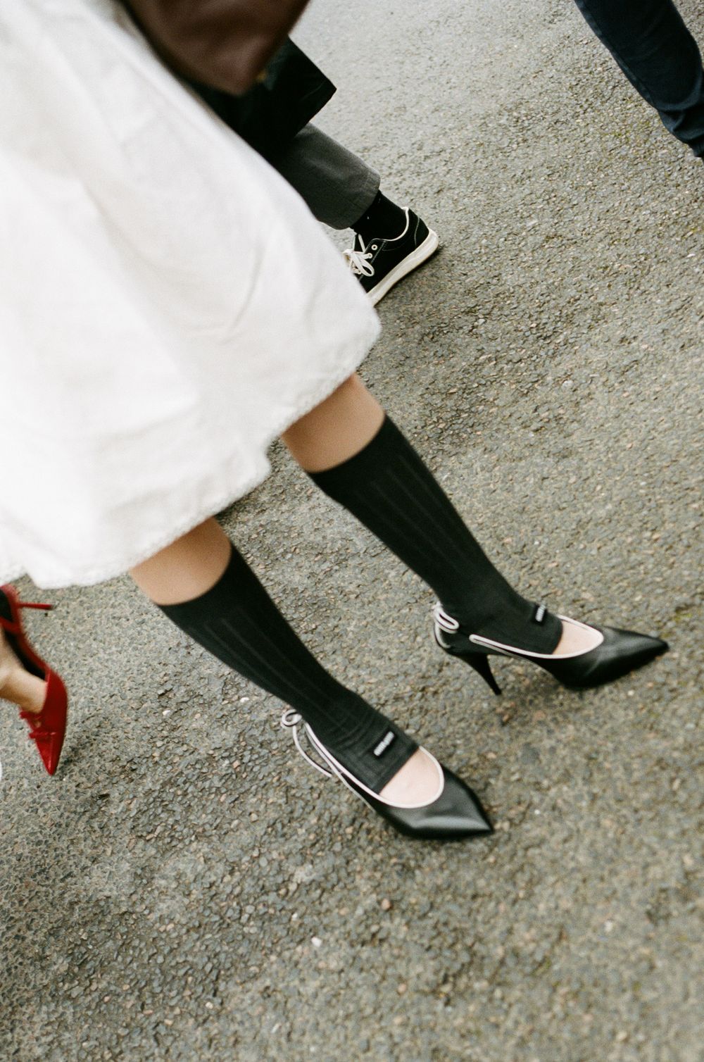 Two pairs of feet wearing black knee-high socks with contrasting white dress and black pointed-toe shoes on concrete.