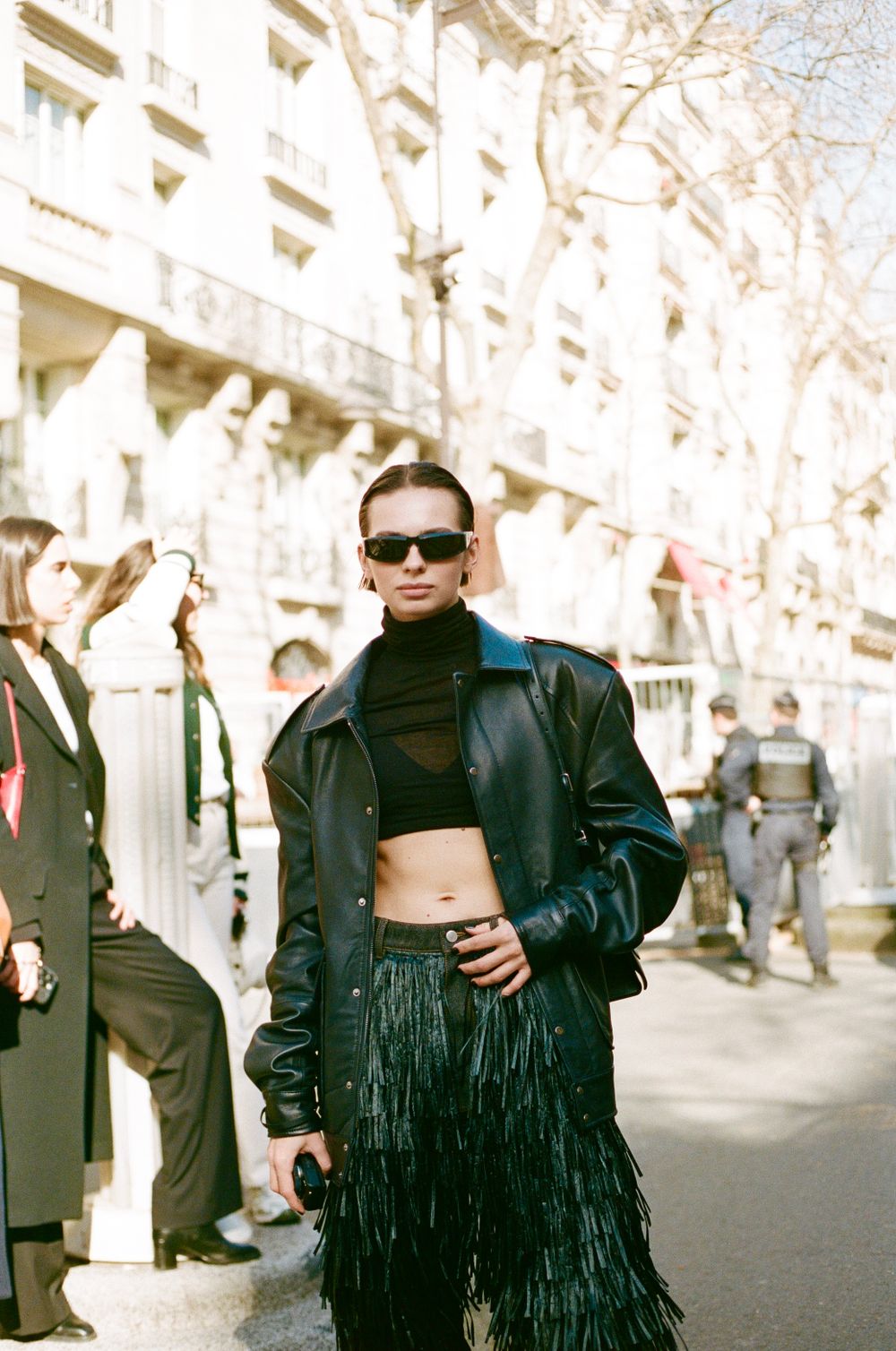 Street style fashion featuring a black leather jacket, cropped turtleneck and fringe skirt worn with sunglasses in Paris.